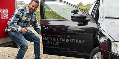 Martins Fahrschule Düsseldorf – Fahrlehrer Martin Wohlgemuth zeigt auf Fahrschulfahrzeug mit Firmenlogo