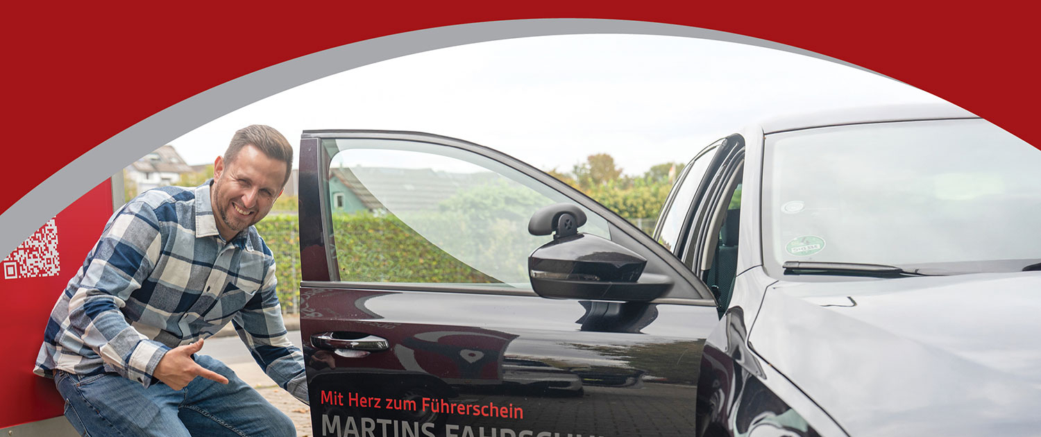 Martins Fahrschule Düsseldorf – Fahrlehrer Martin Wohlgemuth zeigt auf Fahrschulfahrzeug mit Firmenlogo